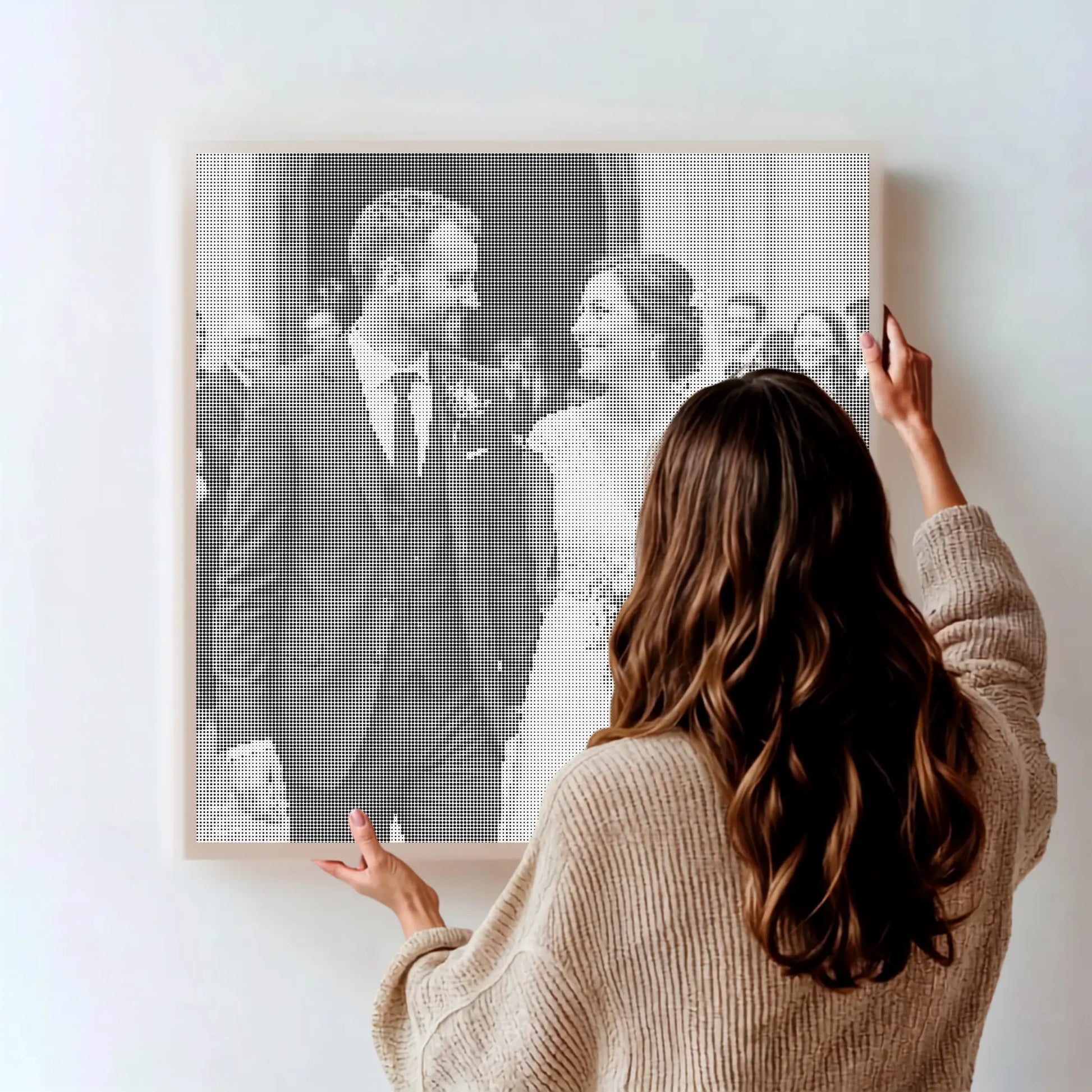 A woman in a beige knit sweater stands with her back to the camera, holding up a framed black-and-white wedding photograph on a light wall. The photo shows a smiling couple walking indoors, and the image is presented as a styled lifestyle shot.